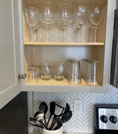 Fully stocked kitchen cabinet with wine glasses, tumblers, and utensils.